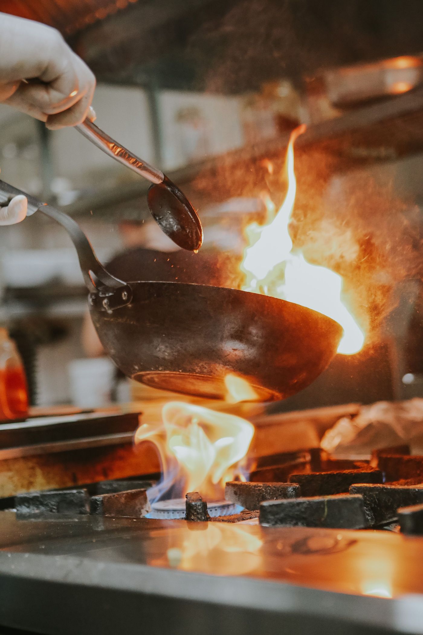 A cook tilting a steel pan over open flame on a restaurant range